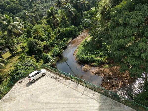 A white car parked on a paved area adjacent to a river, surrounded by lush greenery and a metal railing.
