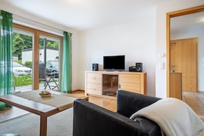 A modern living room with a sofa, a television on a wooden cabinet, and a sliding glass door leading to a parking area.