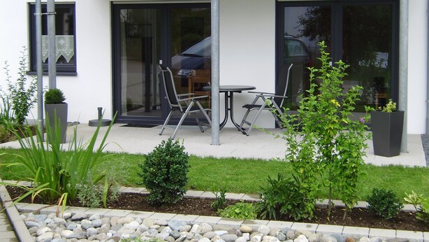 Ein modernes Gebäude mit einem Balkon, einer Terrasse mit Tisch und Stühlen sowie einem Gartenbereich mit Pflanzen und einem Steingarten.