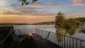A lakeside deck with a fire pit and chairs, overlooking a calm lake with trees and houses in the background. - Waterfront Retreat | Dock, Kayak & Lake Views (Hopkinton)