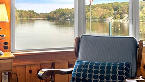 A room with a large window overlooking a lake, a wooden chair with a patterned cushion, and a lamp on a wooden nightstand. - Waterfront Retreat | Dock, Kayak & Lake Views (Hopkinton)