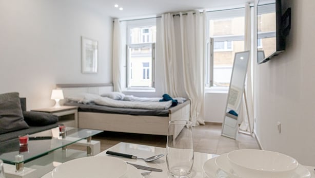 Une chambre d’hôtel moderne comprenant un lit, une table basse en verre, une table à manger avec de l’assiette et des couverts, un téléviseur fixé au mur et une fenêtre avec des rideaux.