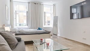 Une chambre moderne avec un lit, une table basse en verre, un téléviseur fixé au mur et une fenêtre avec des rideaux.