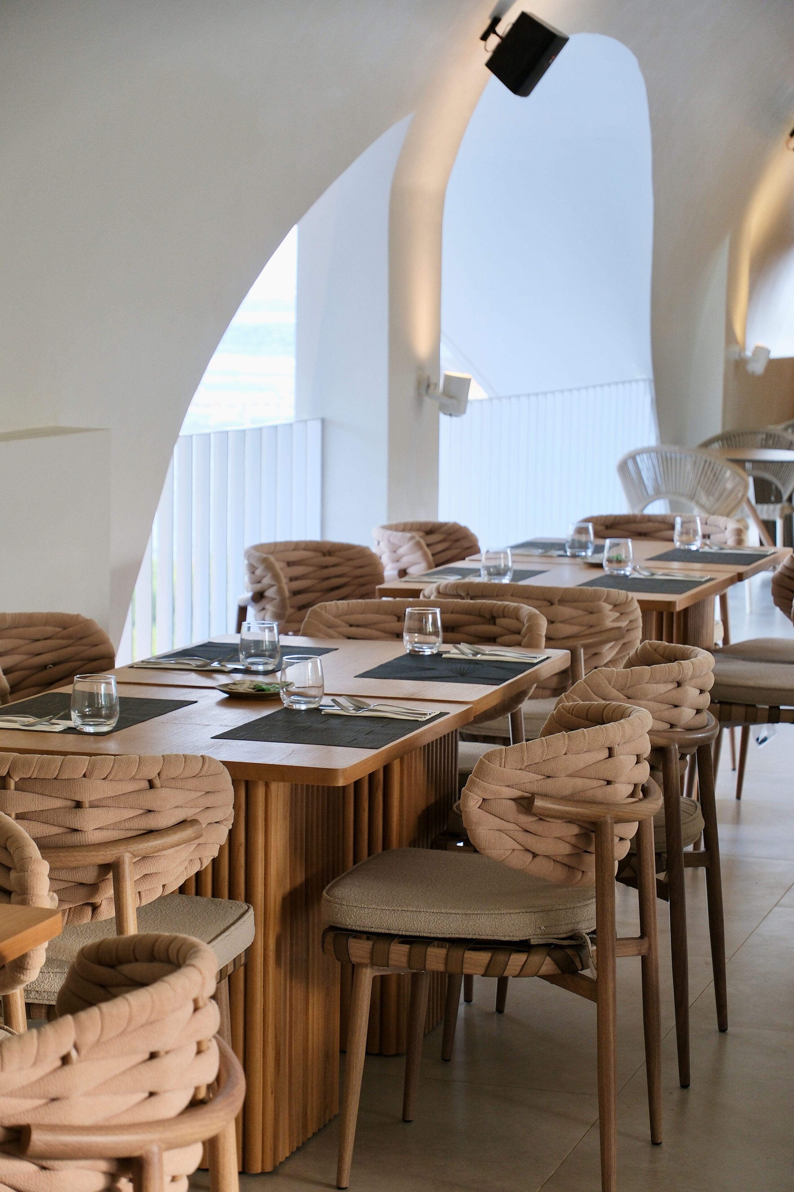 A restaurant with high tables and woven chairs, a modern interior, and a view through an arched window.