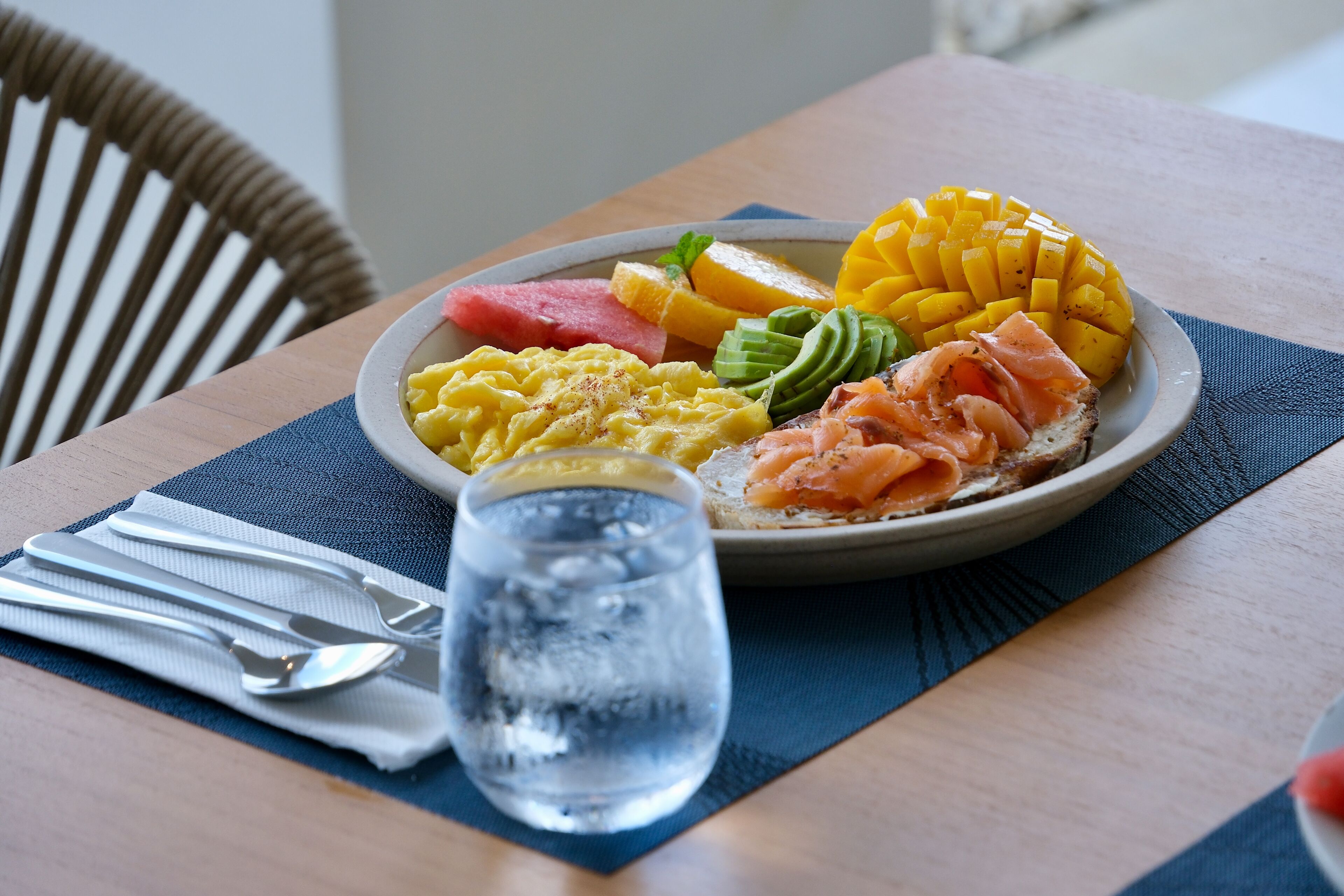 A plate of food with scrambled eggs, avocado, smoked salmon, mango, and watermelon on a table with a water glass and cutlery.