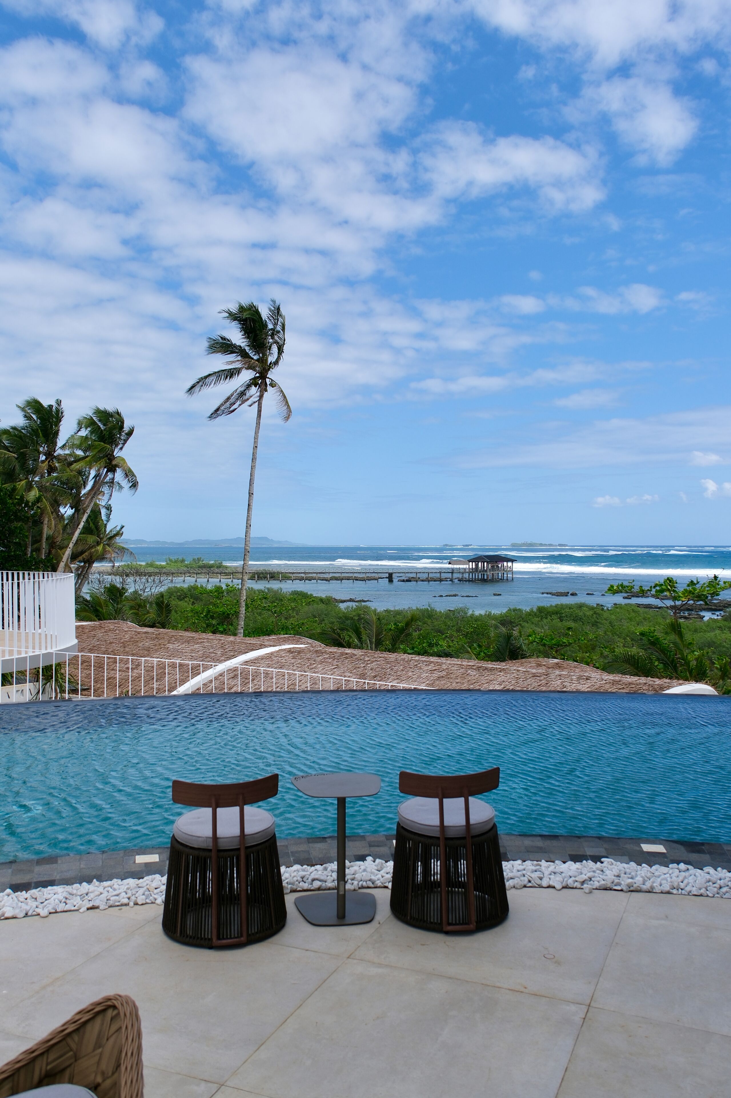 Une piscine à débordement avec deux chaises et une table, entourée de palmiers et offrant une vue sur l’océan.