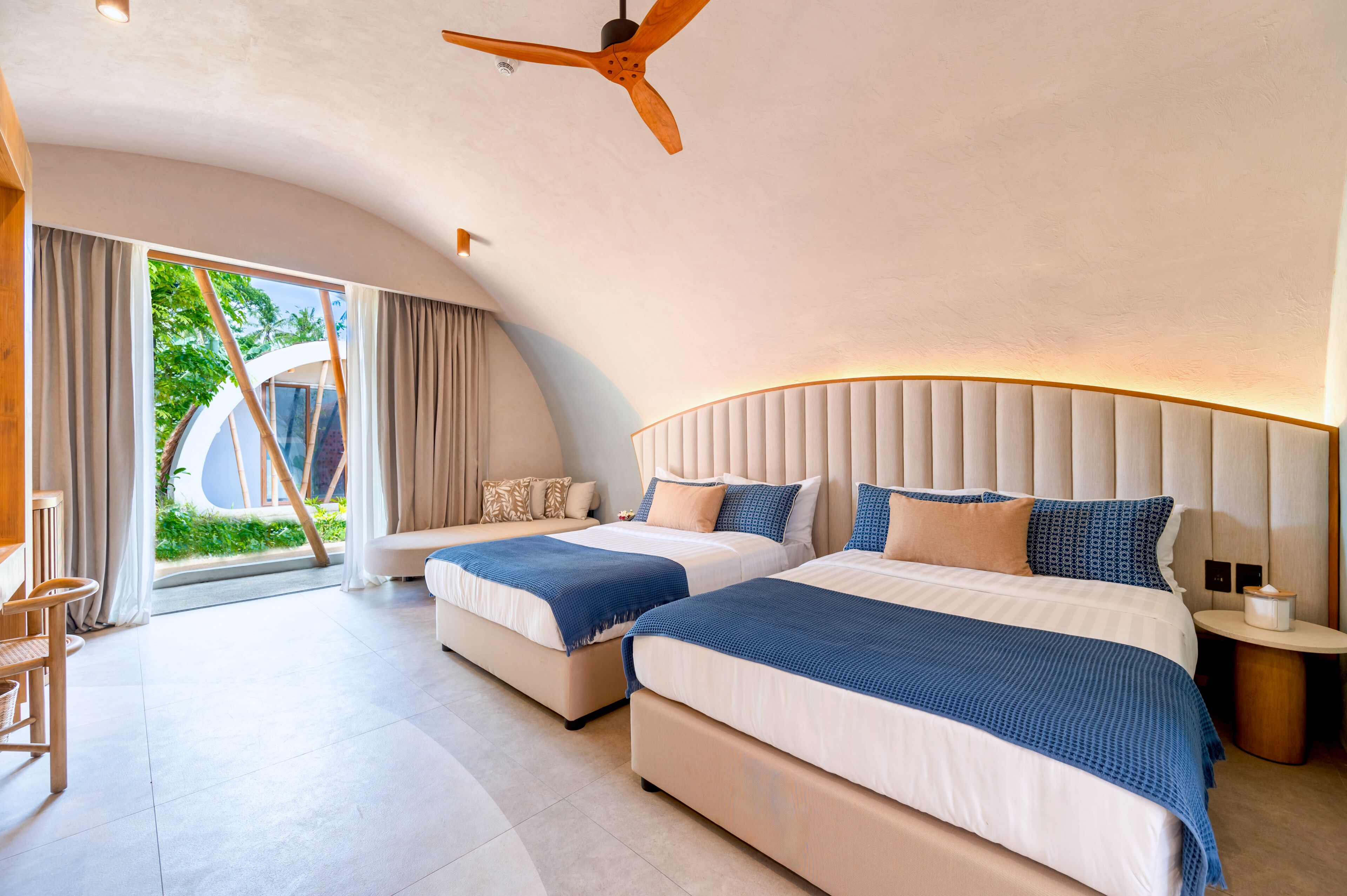 Een moderne hotelkamer met twee bedden, een plafondventilator, een groot raam met gordijnen en uitzicht op groen.