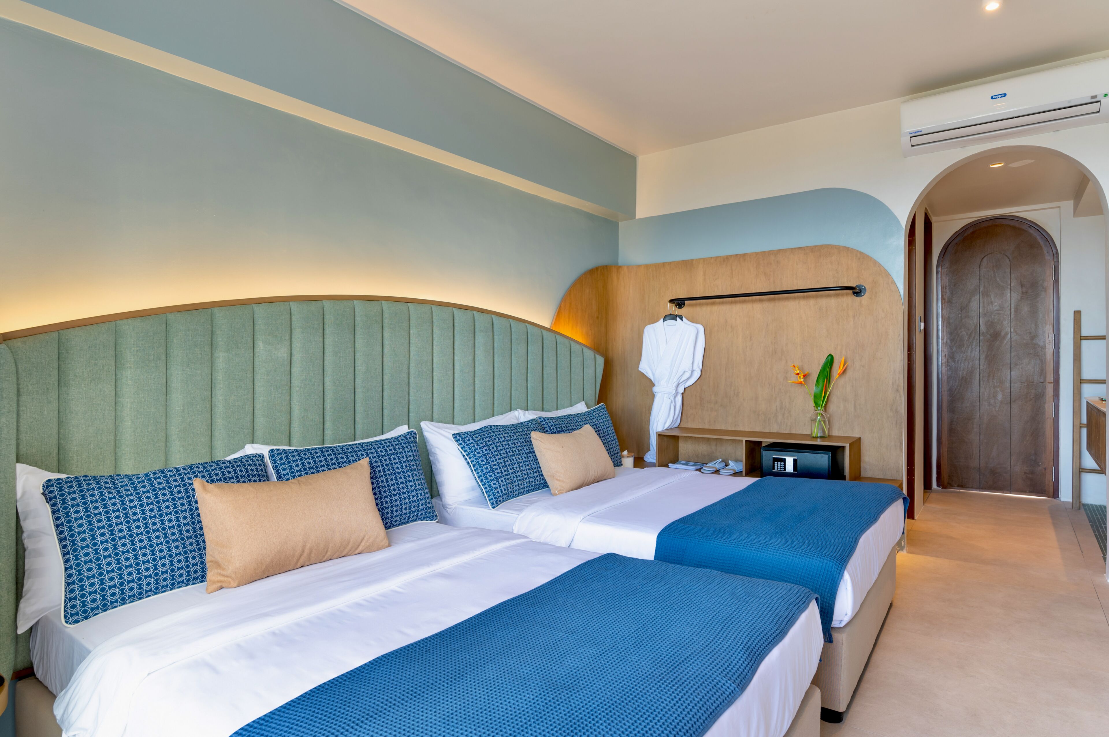 A hotel room with two beds, a curved headboard, a wooden accent wall, a nightstand with a lamp, and a door leading to another room.