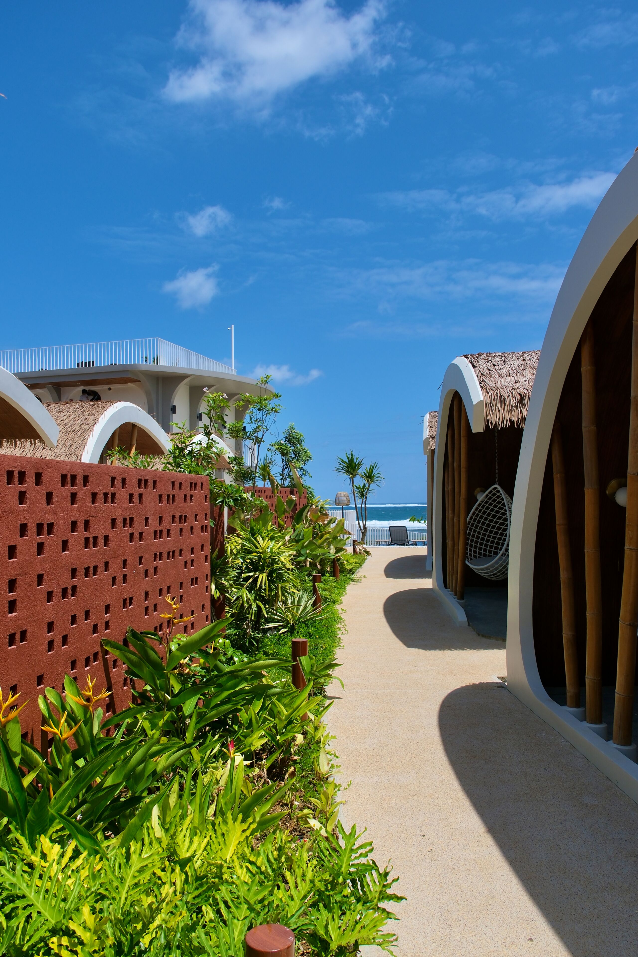 A resort pathway with a thatched-roof structure, a hanging chair, and a view of the ocean.