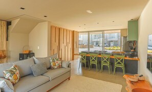 A modern living room with a grey sofa, wooden panel wall, and a kitchen area with bar stools.