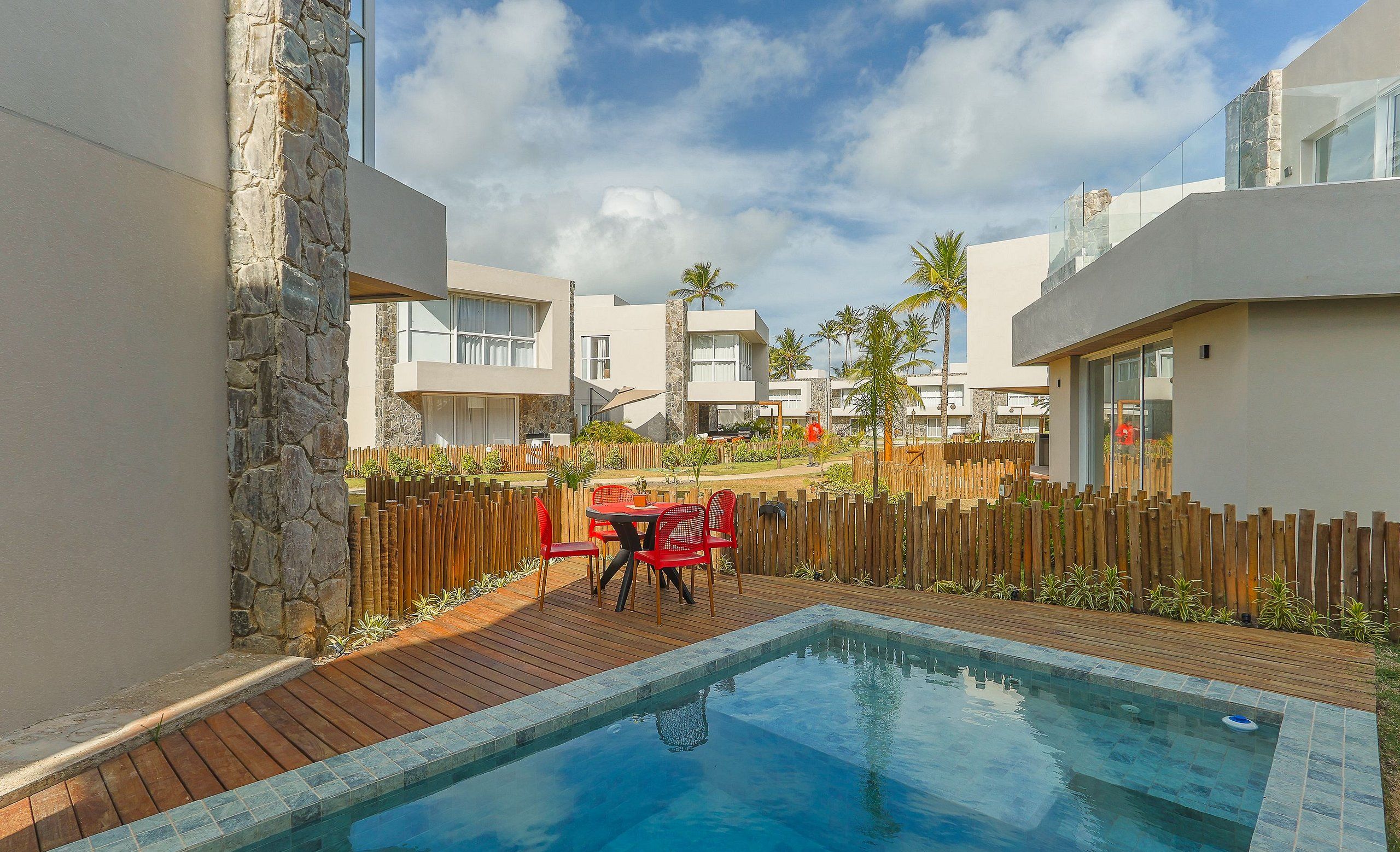 Un área de piscina moderna con terraza de madera, paredes de piedra y una mesa roja con sillas.