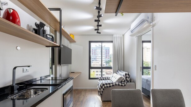 Una cocina moderna con encimeras negras, un lavamanos de acero inoxidable y un hervidor rojo. Se observa una zona de comedor con una mesa de madera y dos sillas. Se ve una cama con una colcha a cuadros a través de una puerta abierta.