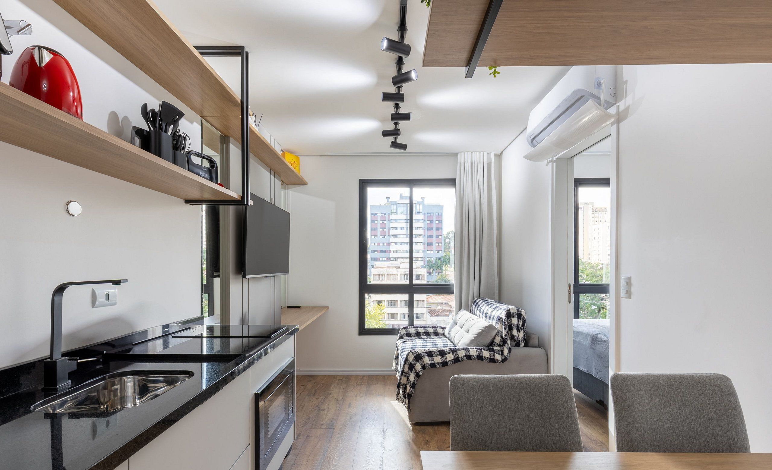 A modern kitchen with black countertops, a stainless steel sink, and a red kettle. There is a dining area with a wooden table and two chairs. A bed with a checkered blanket is visible through an open door.