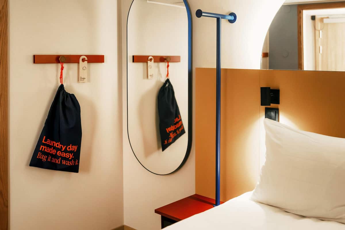 Habitación de hotel con cama, mesita de noche, espejo y un perchero en la pared con una toalla negra y una etiqueta.