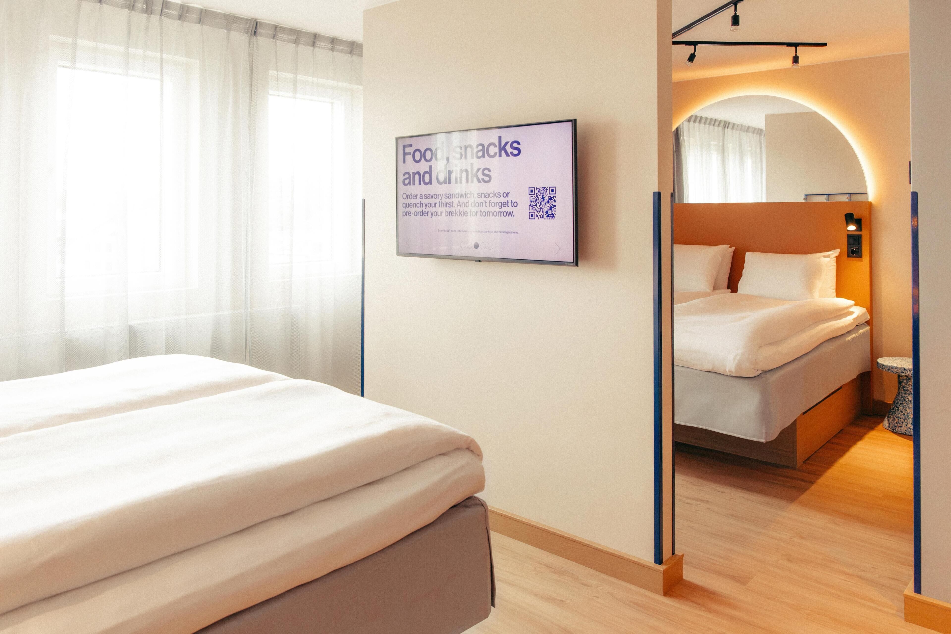 A hotel room with a bed, a bedside table, a mirror, a wall-mounted TV, and a sign about food, snacks, and drinks.