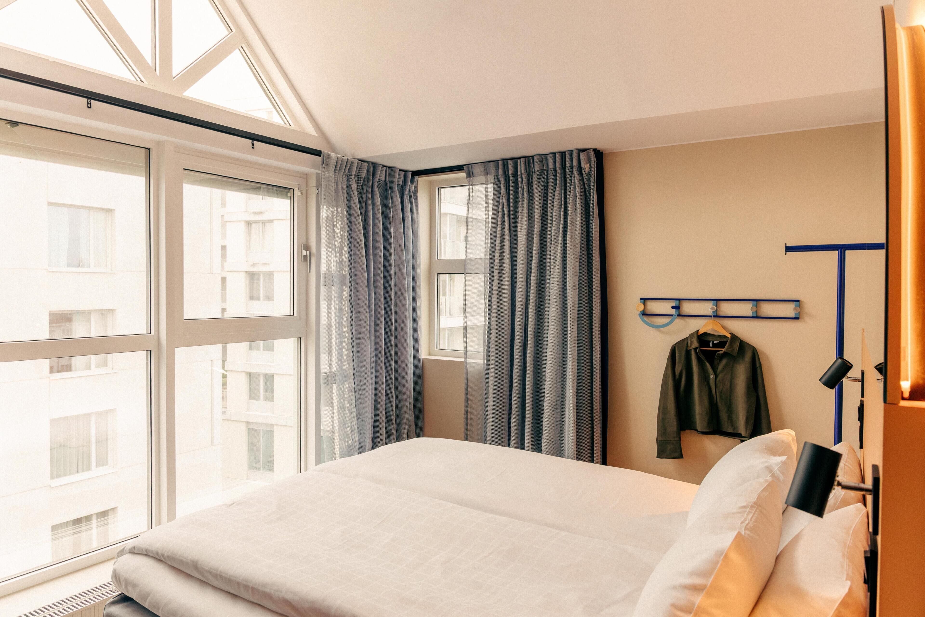 Ein Hotelzimmer mit Bett, großen Fenstern, einem Kleiderständer und einem aufgehängten Mantel.