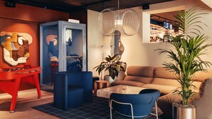Un salon moderne comprenant un canapé, une table basse, une table rouge, une chaise bleue, une plante en pot et un luminaire suspendu original.