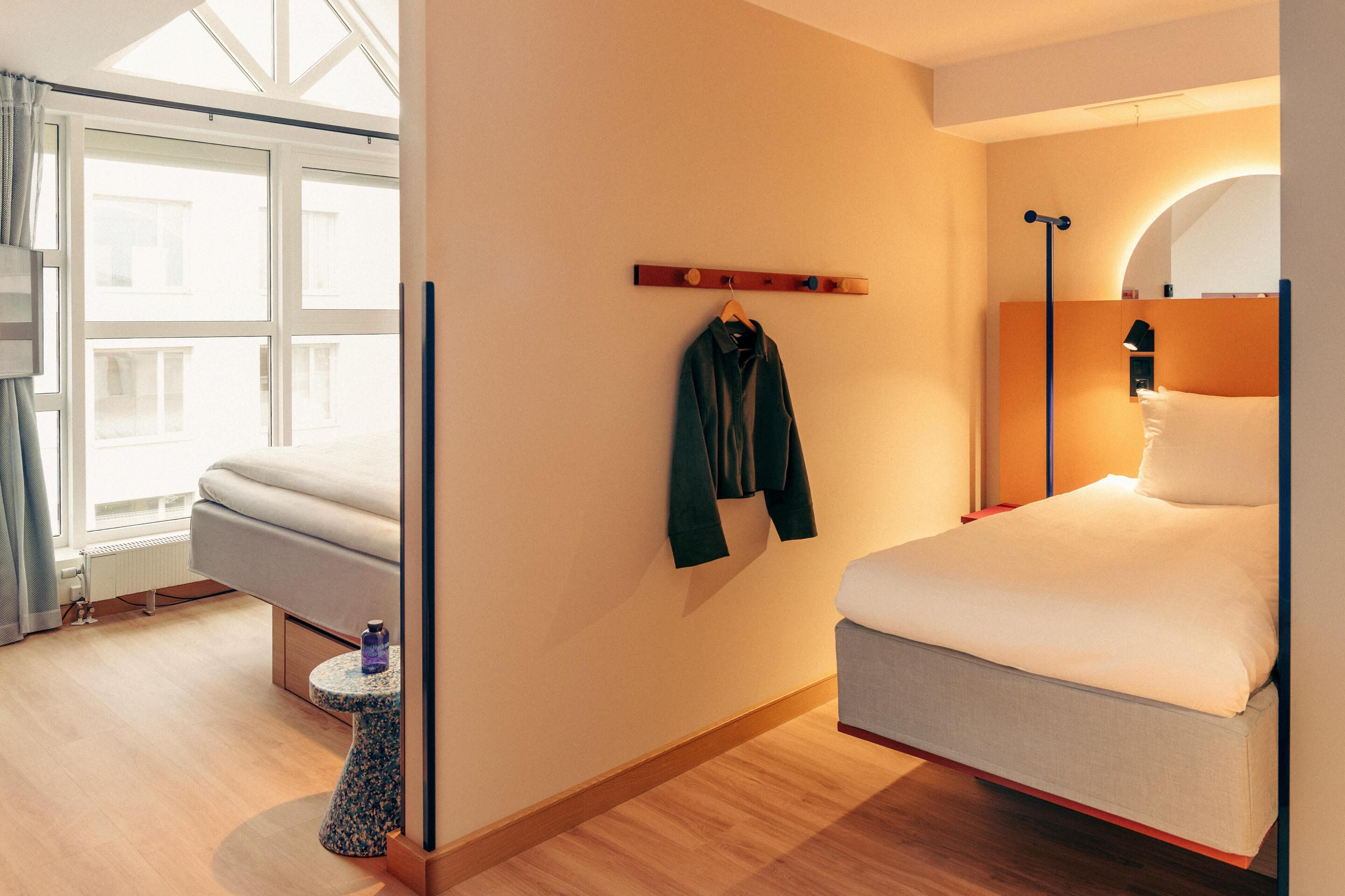 A modern hotel room with a bed, a seating area, a coat rack, and a window.