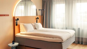 Une chambre d’hôtel moderne, équipée d’un lit, de lampes de chevet, d’une petite table ronde et d’une grande fenêtre avec des rideaux.