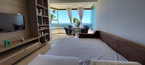 A hotel room with a bed, a TV mounted on the wall, a bookshelf, a balcony with a table and chairs, and a view of the ocean.