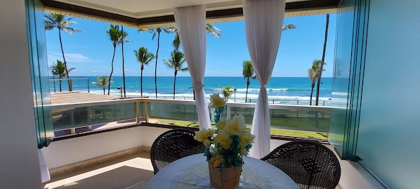 A balcony with a view of the ocean, palm trees, and a table with flowers.