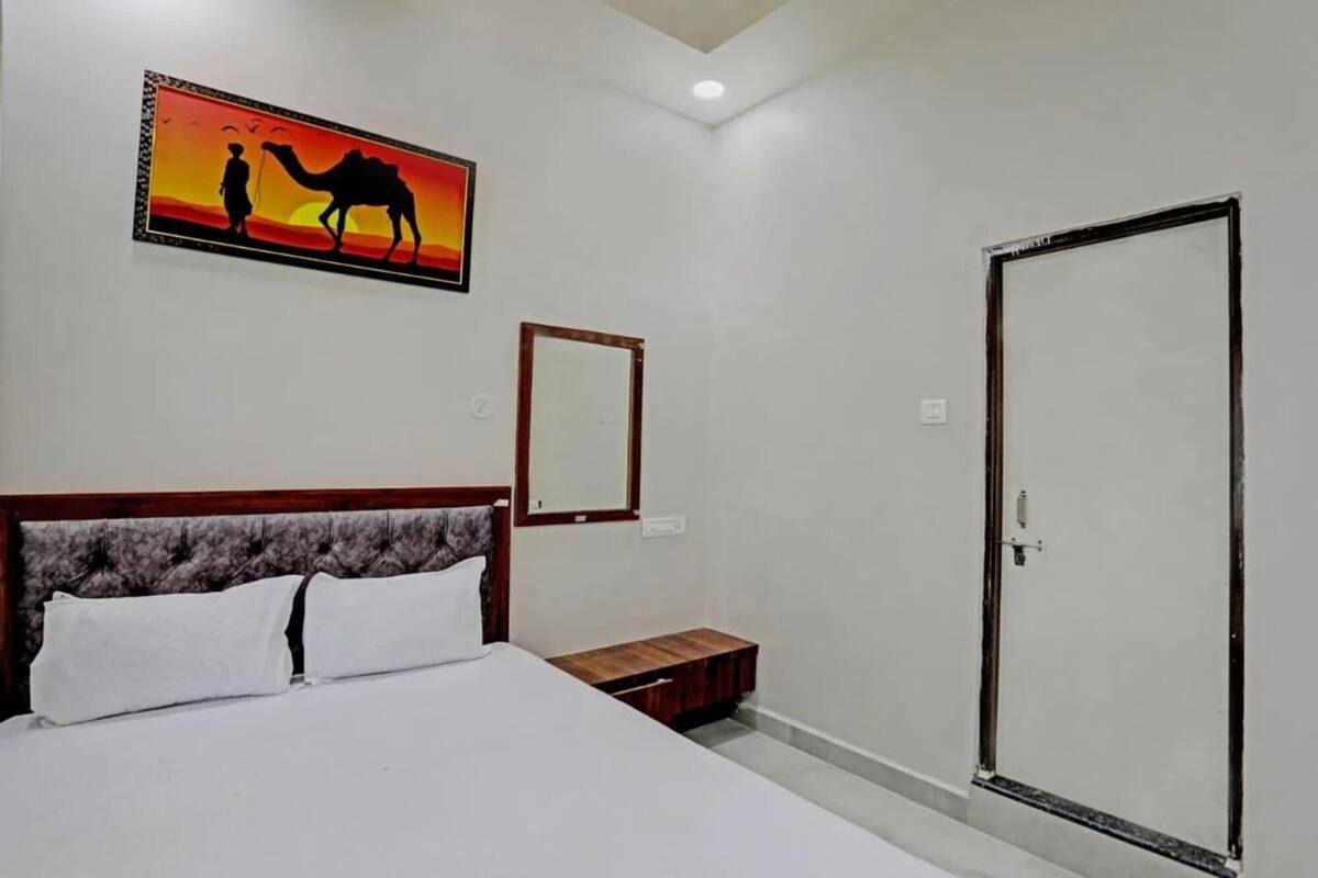 Een slaapkamer met een bed, nachtkastje, kledingkast, spiegel en een muur met een zonsondergangschilderij.