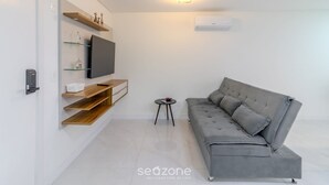 Ein modernes Wohnzimmer mit grauem Sofa, einem Flachbildfernseher, Holzregalen und einem kleinen Beistelltisch.