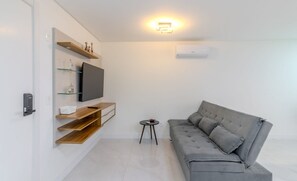 Uma sala de estar moderna com um sofá cinza, uma TV de tela plana, prateleiras de madeira e uma pequena mesa lateral.