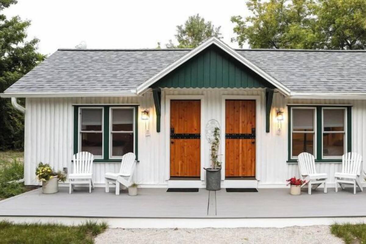 A small, single-story building with a gabled roof, two front doors, and a porch with white chairs.