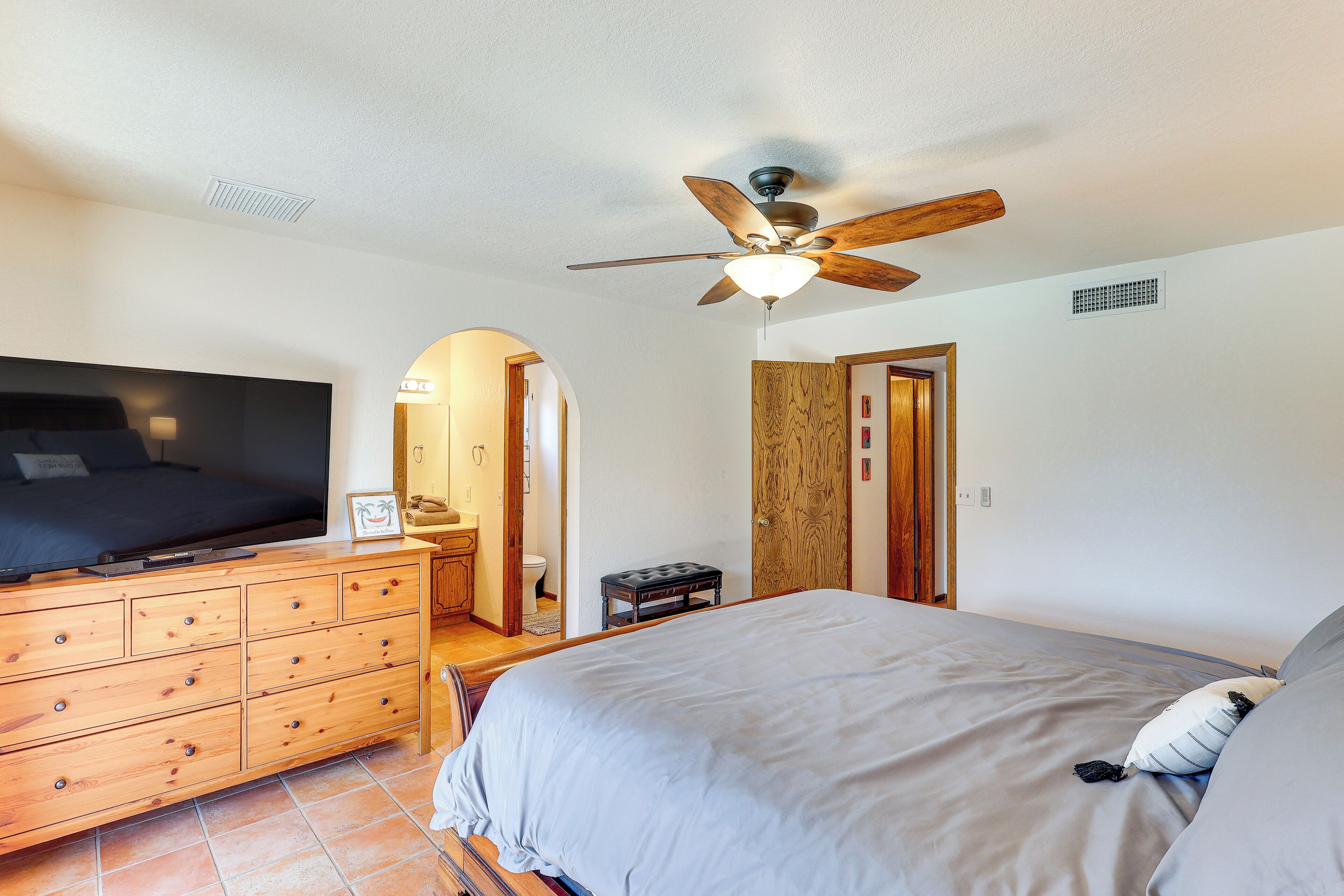 A bedroom with a bed, a wooden dresser, a ceiling fan, and a bathroom visible through an archway.