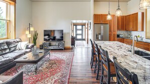 A modern living room with a leather sofa, a coffee table, and a TV. The kitchen features wooden cabinets and a marble island.