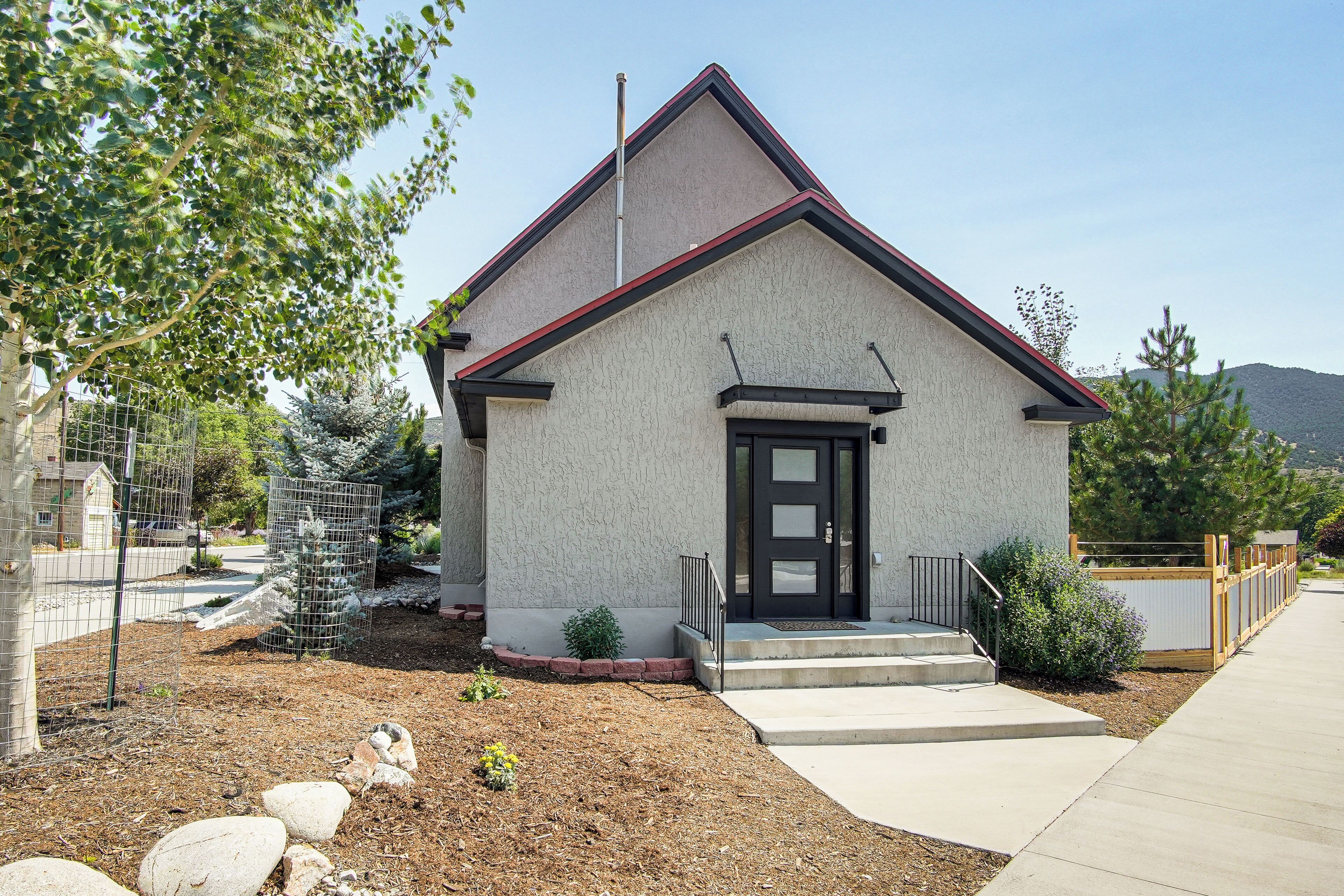 A small, single-story building with a red roof and a black door, surrounded by a landscaped garden with rocks and a small tree.