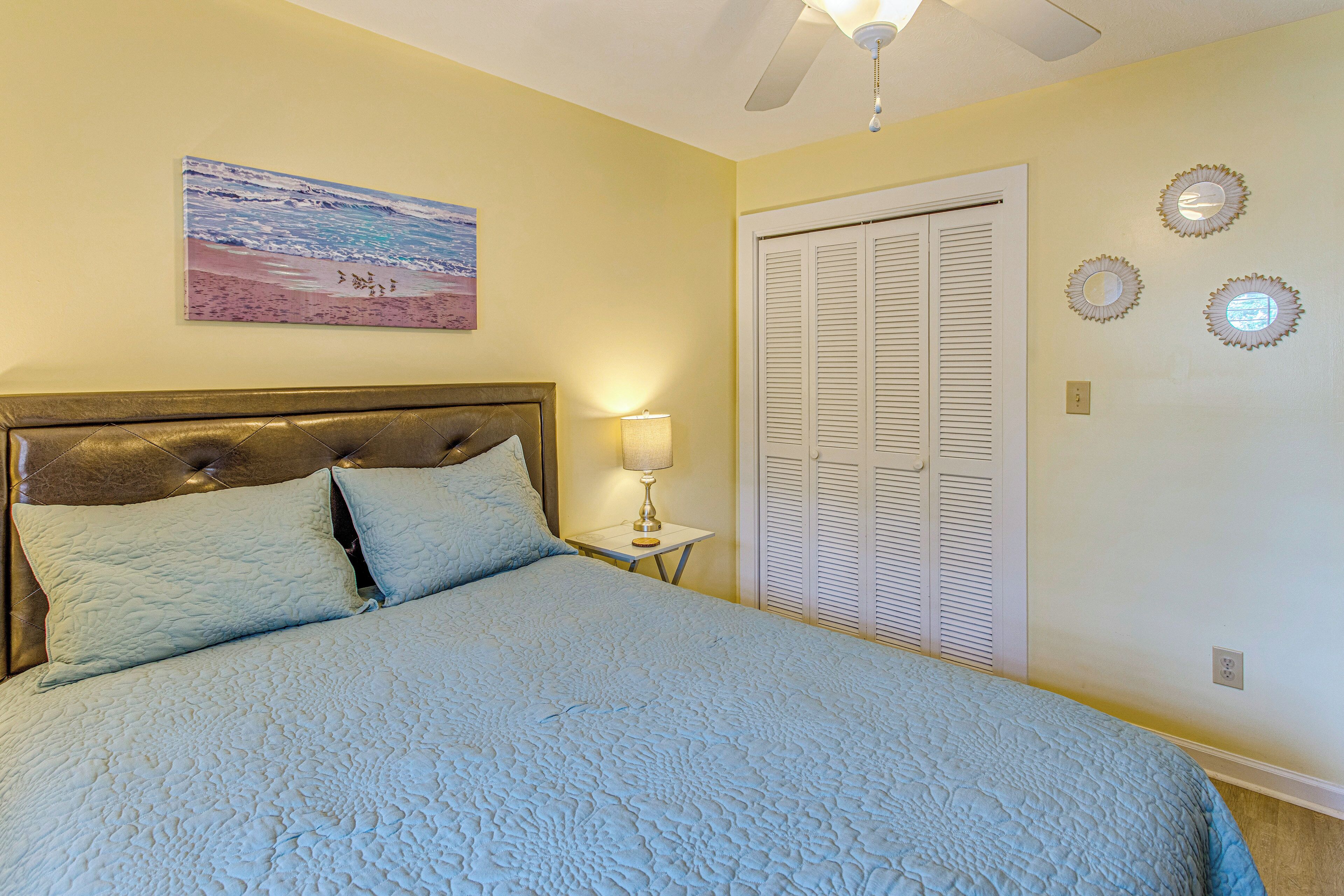 A bedroom with a bed, a nightstand, a lamp, a ceiling fan, a beach painting, and a closet with louvered doors.