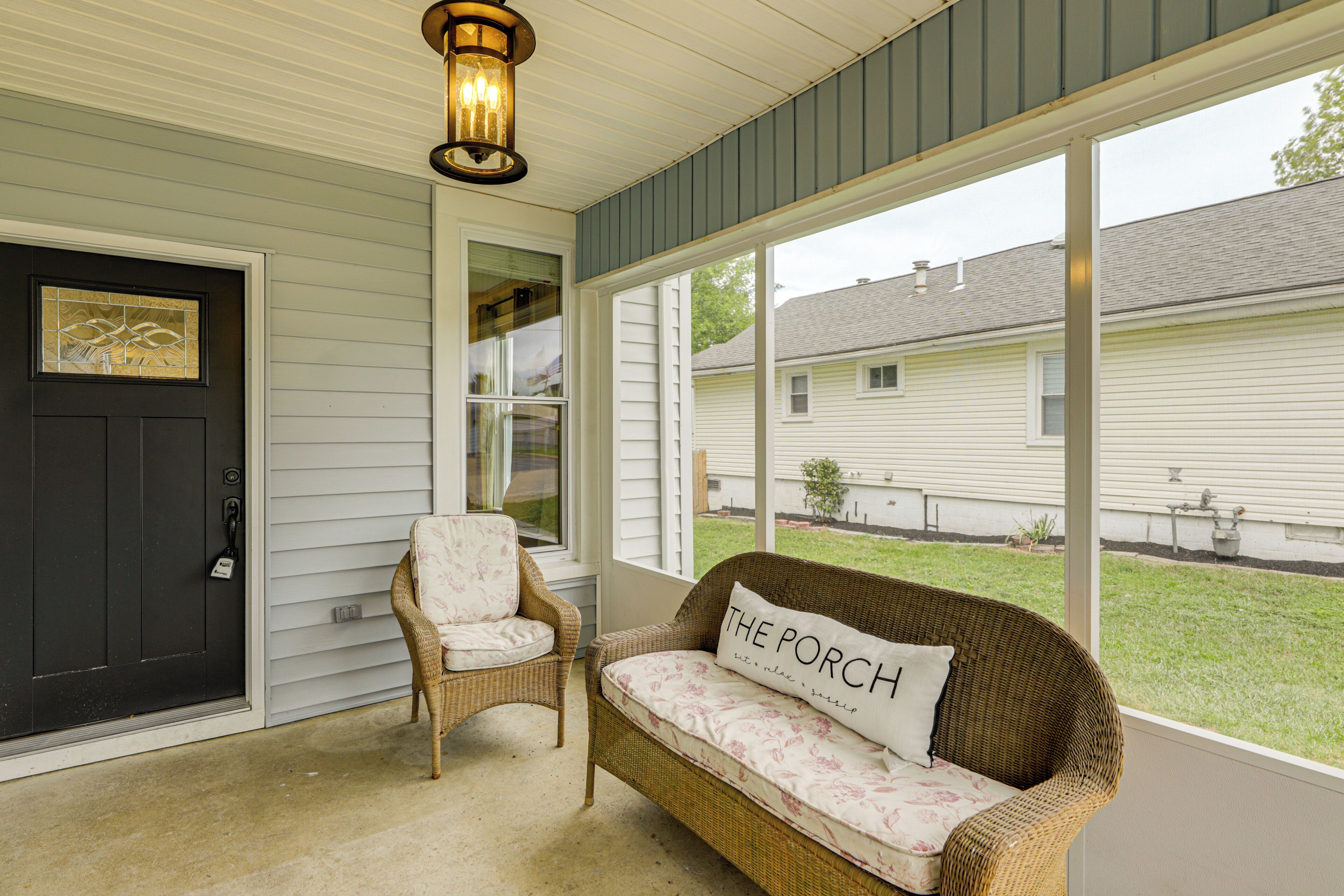 A porch with wicker furniture and a decorative pillow, a door with a window, and a view of a house and lawn.