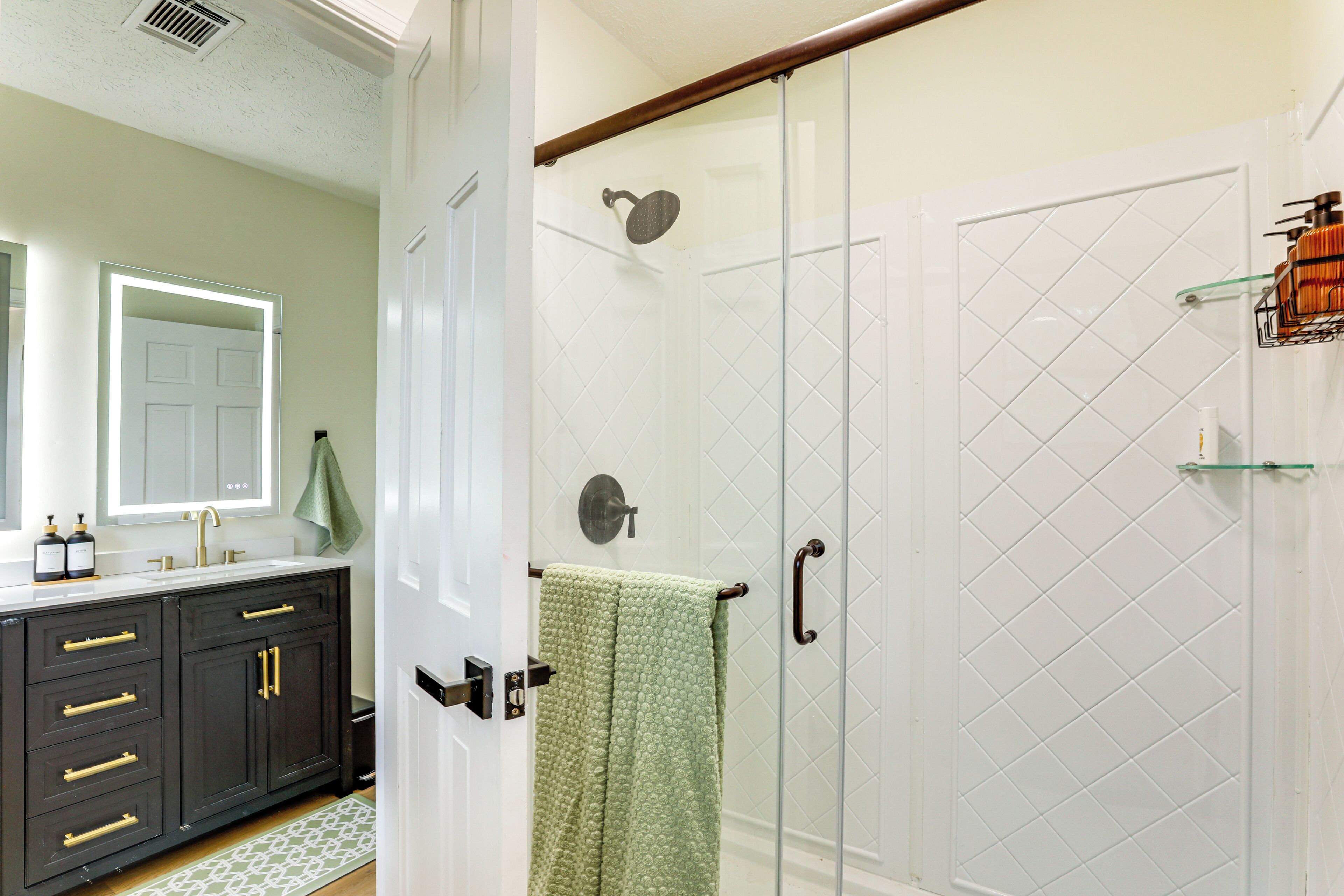 A bathroom with a glass shower enclosure, a dark wooden vanity with gold handles, and a green towel hanging on the door.