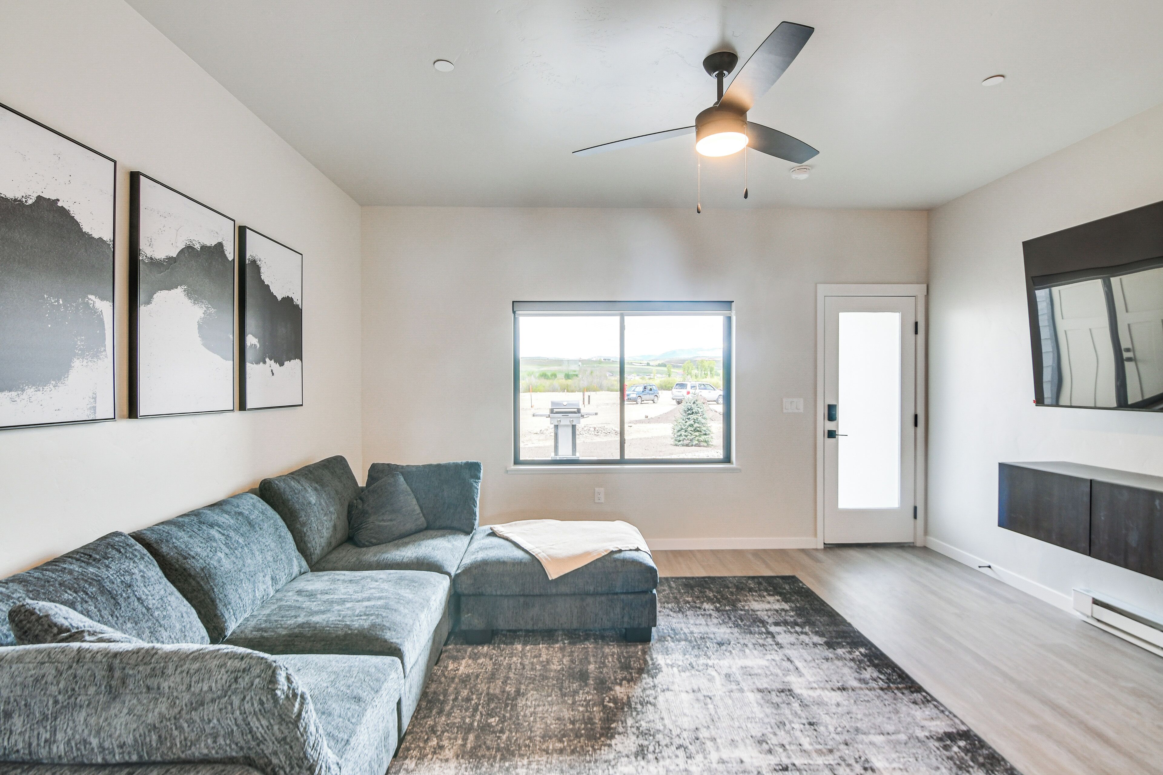 A modern living room with a sectional sofa, a ceiling fan, a large window with a view, and artwork on the wall.
