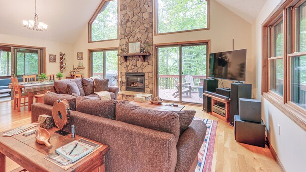 A spacious living room with a stone fireplace, large windows, a television, and a cozy seating area. - Great Cacapon Mtn Cabin w/ Private Hot Tub + Views (Great Cacapon)