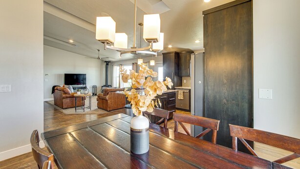 A spacious dining area with a wooden table, chairs, and a vase of flowers. There is a kitchen in the background with dark cabinets and a large door. - Panoramic Views: Spacious Home in Cedar City! (Cedar City)