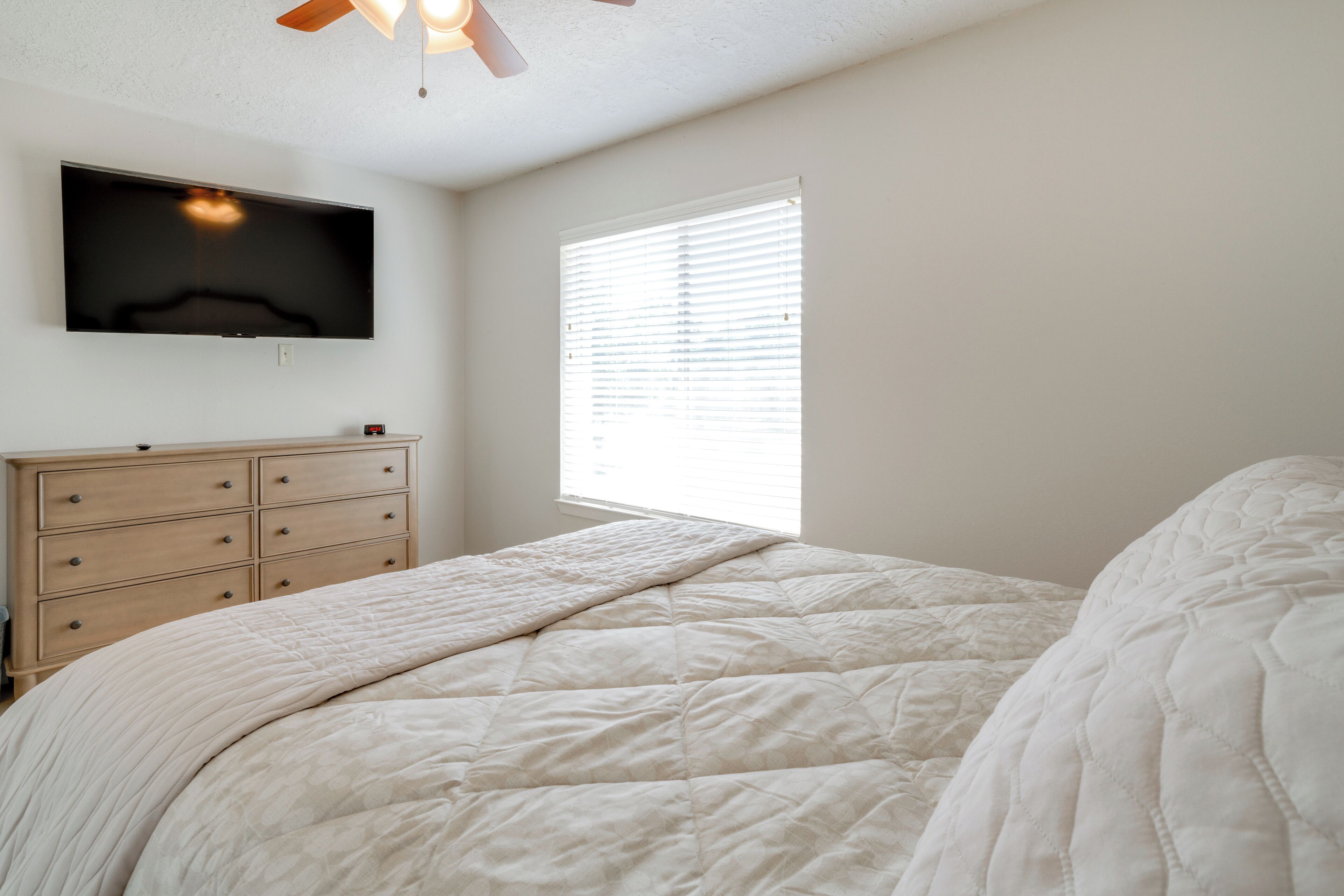 A bedroom with a bed, a dresser, a TV mounted on the wall, and a window with blinds.