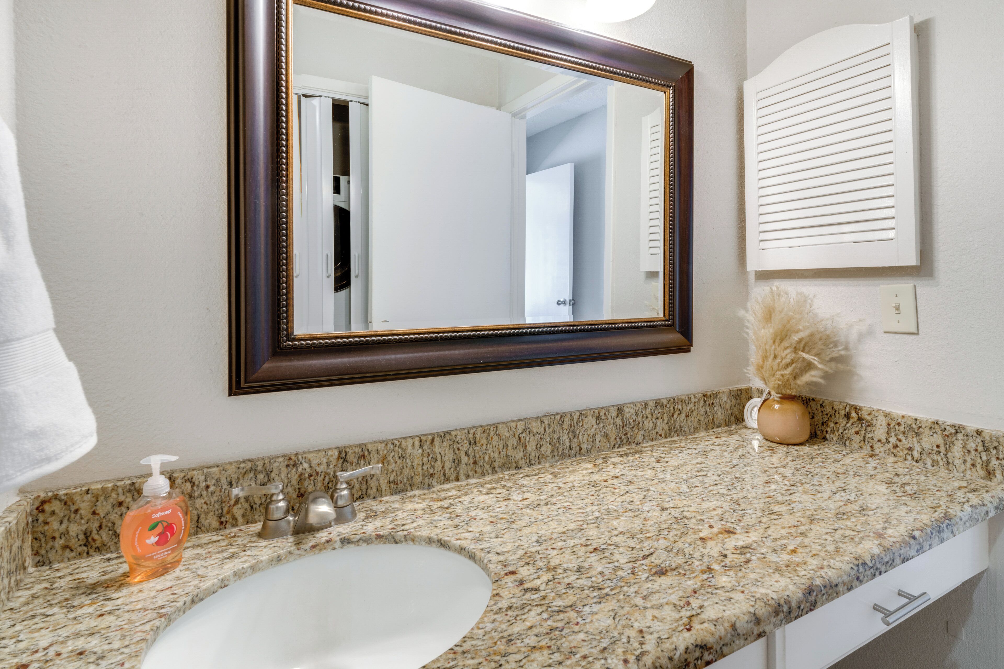 A bathroom with a granite countertop, a white sink, a mirror with a wooden frame, a bottle of hand soap, and a vase with dried flowers.