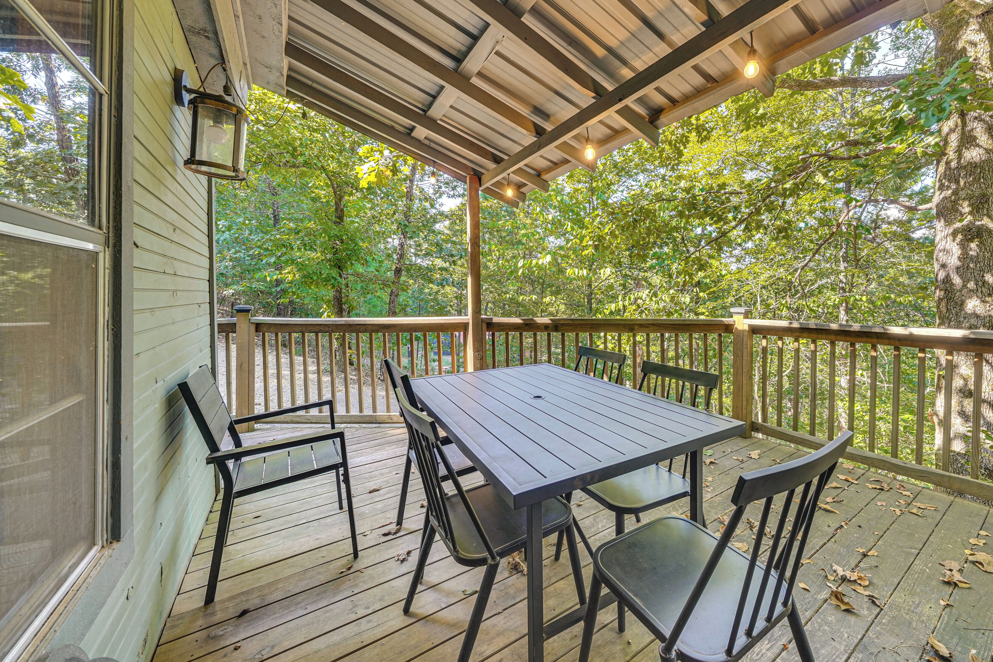 A covered deck with a table and chairs, surrounded by a wooden railing, overlooking a wooded area.