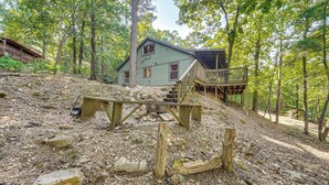 A rustic cabin with a wooden deck and stairs leading to a second level, surrounded by a forested area with a rocky ground. - 3 Mi to Beavers Bend State Park: Wooded Hideaway! (Broken Bow)