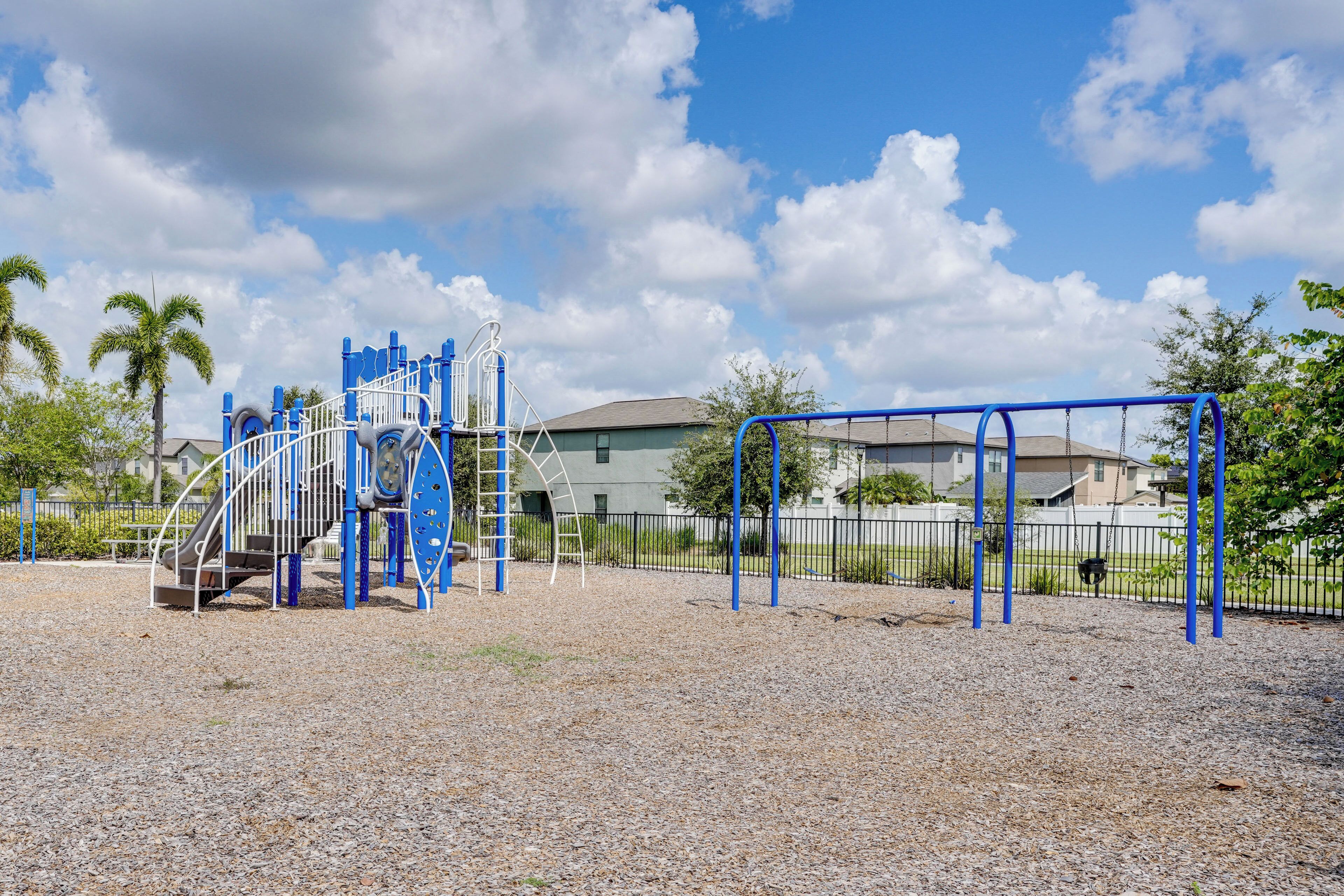 A playground with climbing structures and swings, surrounded by a fence and palm trees.