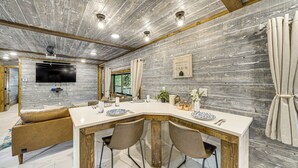 A modern kitchen with a wooden ceiling, a large island with chairs, a TV mounted on the wall, and a sign on the wall. - Hike, Fish & Unwind: Cabin w/ Yard in Broken Bow! (Broken Bow)