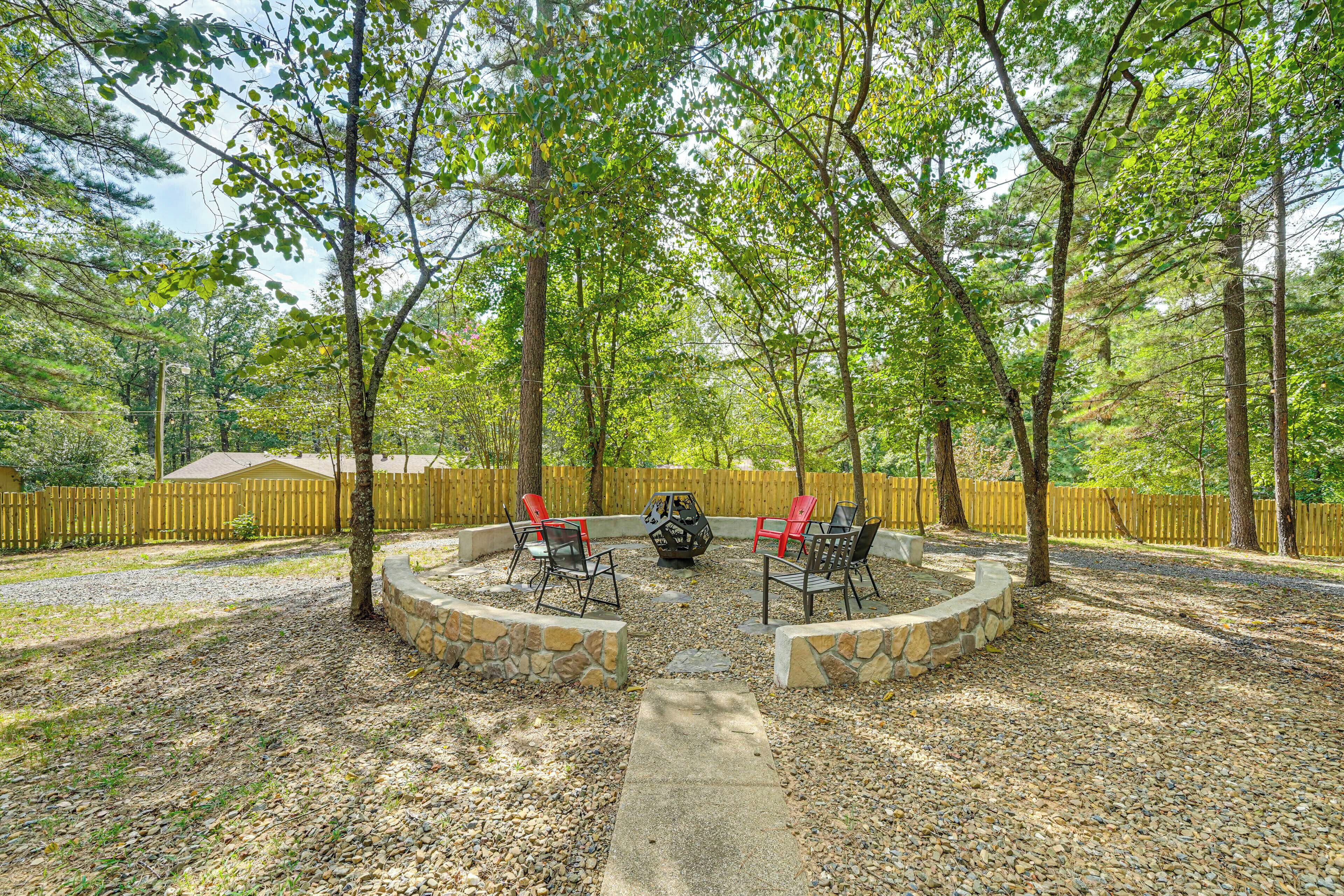 A circular stone fire pit with red and black chairs, surrounded by a stone wall, set in a wooded area with a wooden fence in the background.