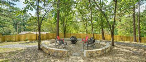 A circular stone fire pit with red and black chairs, surrounded by a stone wall, set in a wooded area with a wooden fence in the background.