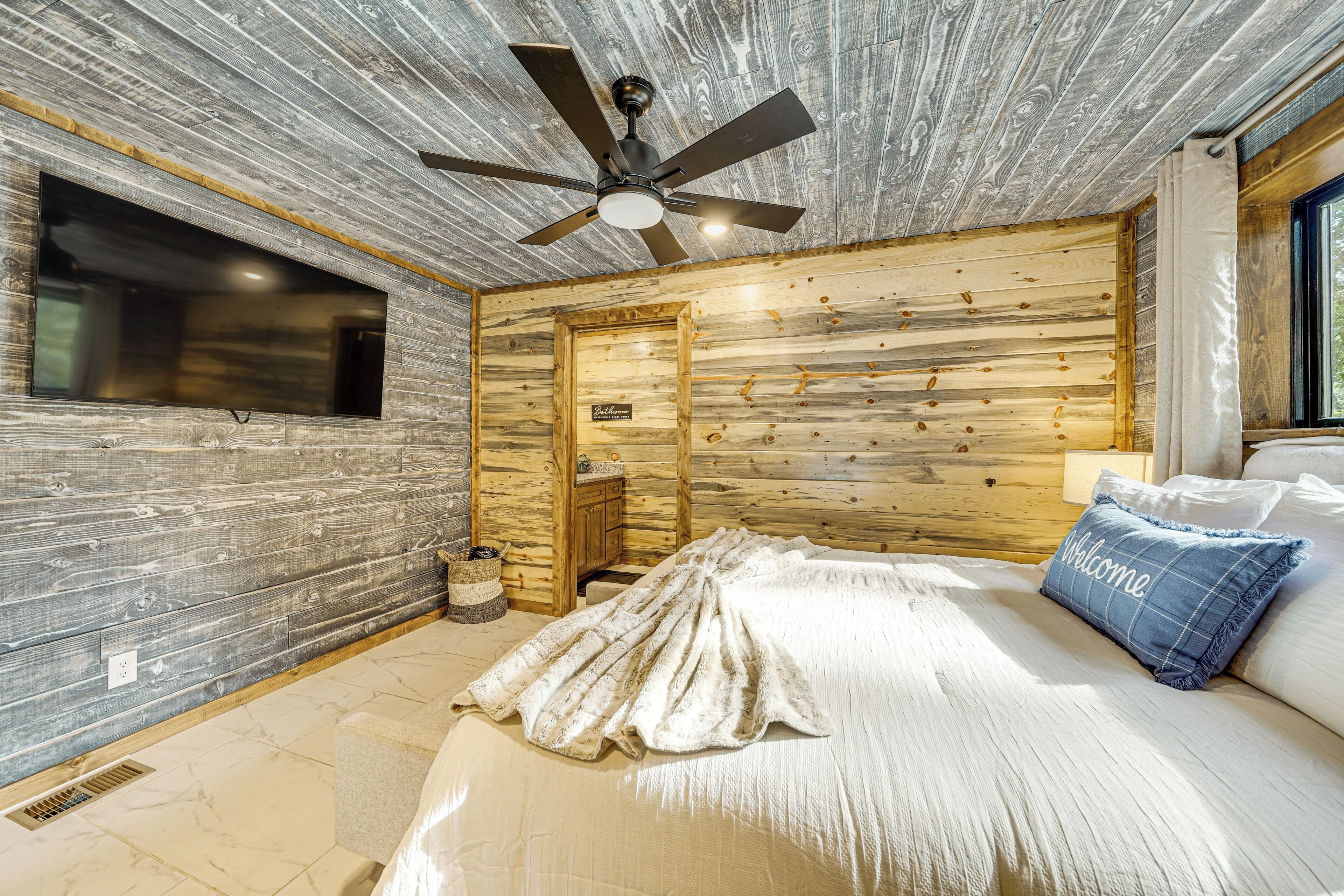 A bedroom with a wooden ceiling, a ceiling fan, a bed with a white comforter and a blue pillow, and a window with curtains.