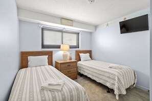 Une chambre avec deux lits, une table de chevet avec une lampe, un téléviseur fixé au mur et une fenêtre avec des stores.