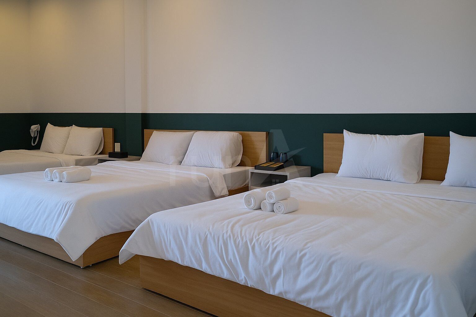A hotel room with three single beds, each with white linens and pillows, and a wooden headboard. There are two bedside tables with lamps and a phone.