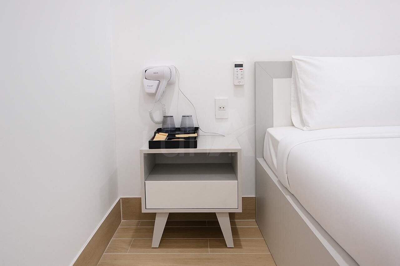Une chambre d’hôtel avec un lit blanc, une table de chevet blanche équipée d’un sèche-cheveux et d’une petite lampe, ainsi qu’une prise électrique au mur.